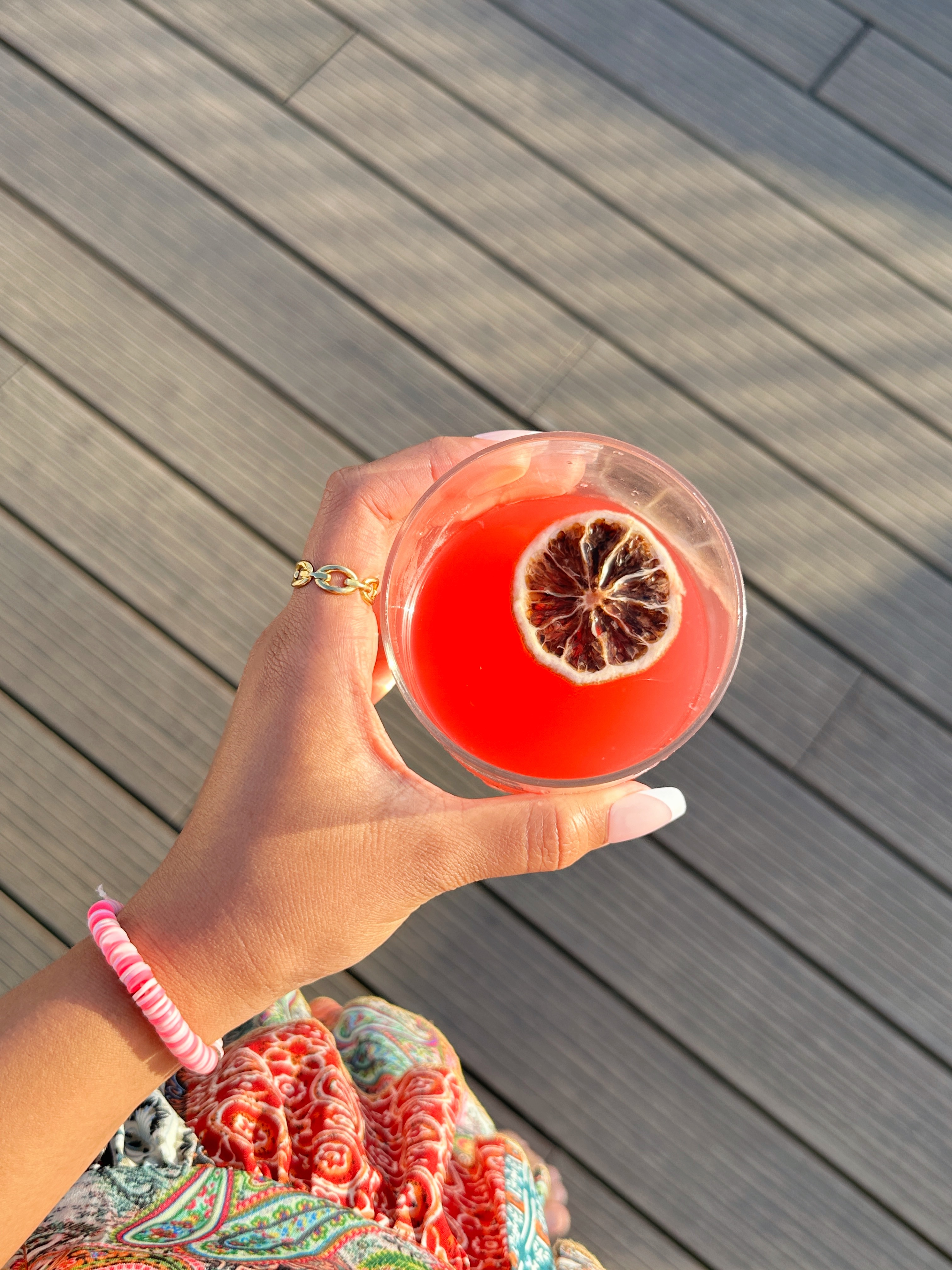 Hand holding a blood orange cocktail with gold jewellery, wooden deck in the background